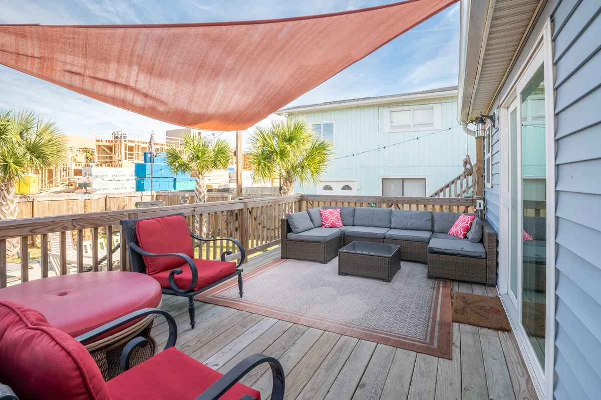 The image shows a vibrant outdoor living space at a single-family home in Carolina Beach, featuring comfortable seating beneath a striking red shade sail, surrounded by swaying palm trees. The area is perfect for relaxing or socializing, with inviting sunset views and ample room for barbecues or stargazing.