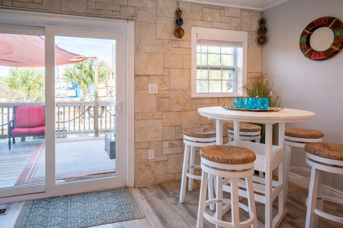 A cheerful interior space featuring a round table with stools, a prominent Tiki bar sign, and sliding glass doors opening to the outdoor deck. The layout suggests a lively spot for casual dining, socializing, and easy transitions between indoor and outdoor spaces.