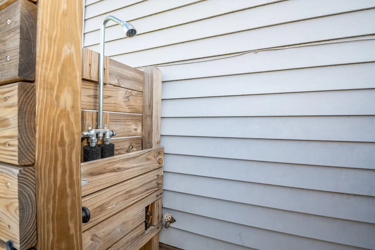 An inviting outdoor shower with a wooden structure and metal showerhead, designed for easy post-beach rinsing. The shower appears approachable and is set in a private area, making it practical for washing off sand and saltwater after beach outings, with a low faucet visible for pets.