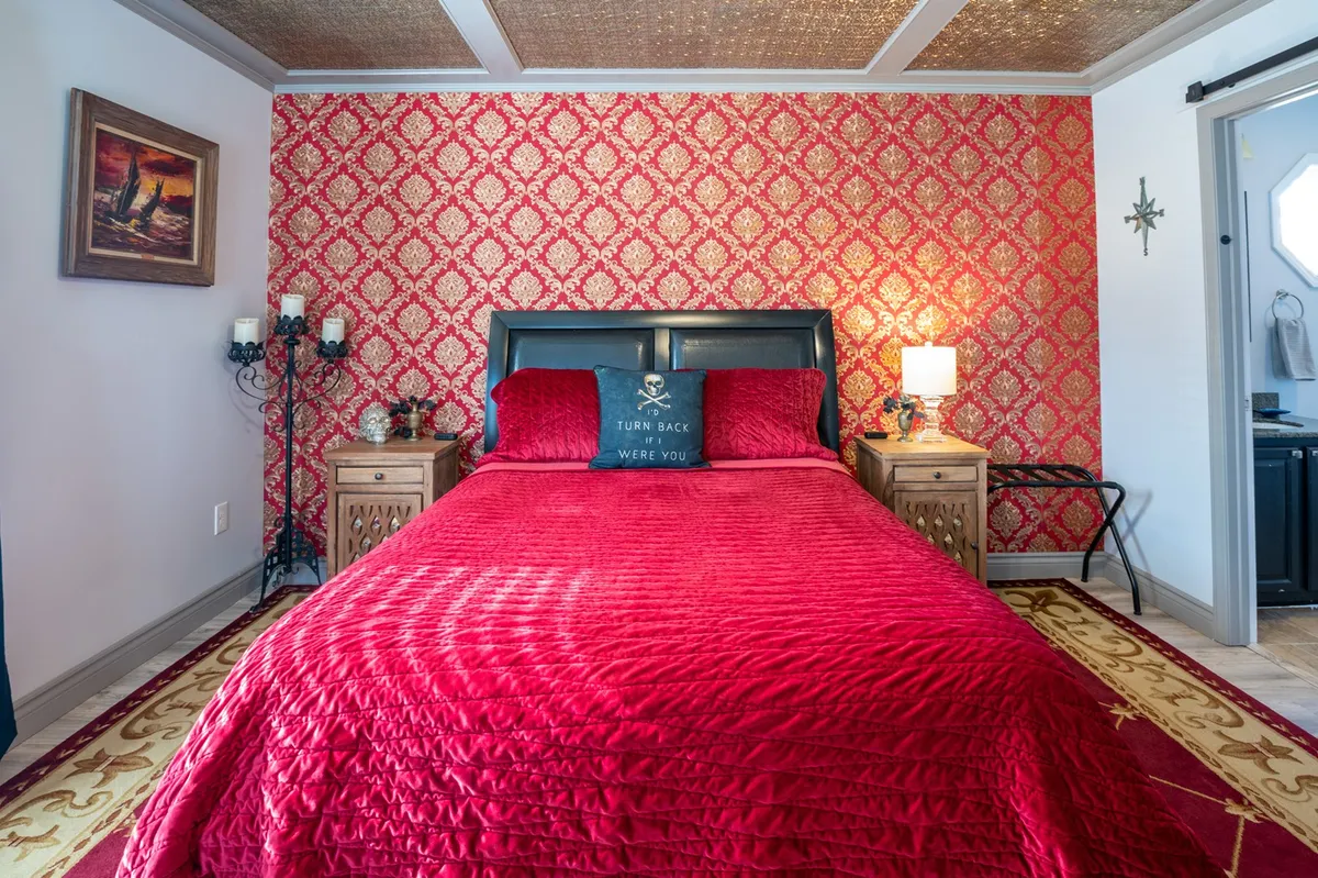 A richly themed bedroom with a bed adorned in red bedding, ornate wallpaper, decorative wall art, nightstands with lamps, and a pirate-themed pillow. The room has playful yet elegant decor, evoking a sense of adventure and luxury.