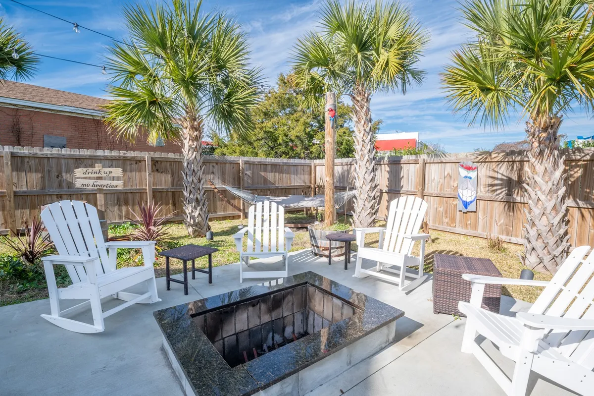An inviting backyard setup with a fire pit surrounded by Adirondack chairs, palm trees, and a hammock nearby. The relaxed, social space encourages gathering around the fire for conversations, stargazing, or unwinding in the hammock under the trees.