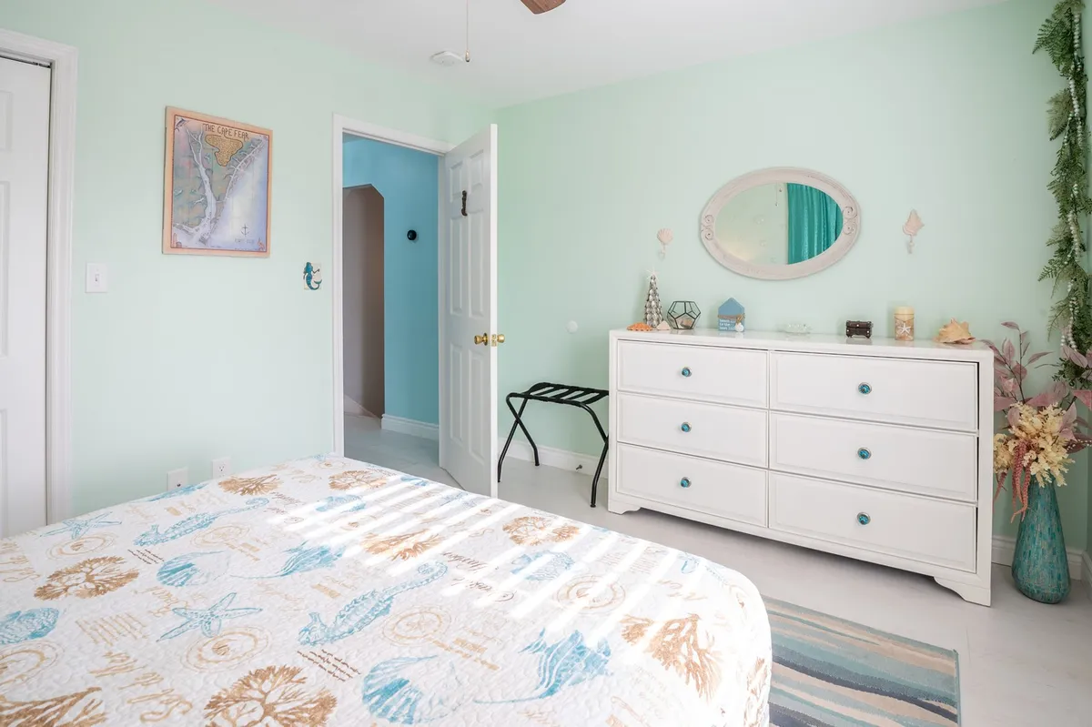 A serene bedroom featuring a bed with a quilt, a dressing table, a world map on the wall, and decorative plants. The space is bathed in natural light and radiates a calming, coastal ambiance.