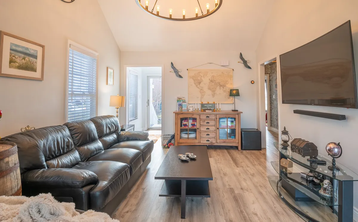 A spacious living room featuring a plush black leather sofa positioned across from a large flat-screen TV. The room also includes a wooden cabinet adorned with a vintage map, subtle nautical accents, and a window letting in soft natural light, creating a welcoming and communal atmosphere.