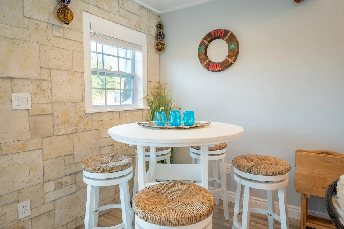 A bright, playful interior scene featuring a 'tiki bar' sign above a cozy bar setup with woven stools, blue glassware, and a round white table. Sunlight pours through the windows, inviting guests to gather and enjoy drinks in a cheerful setting.