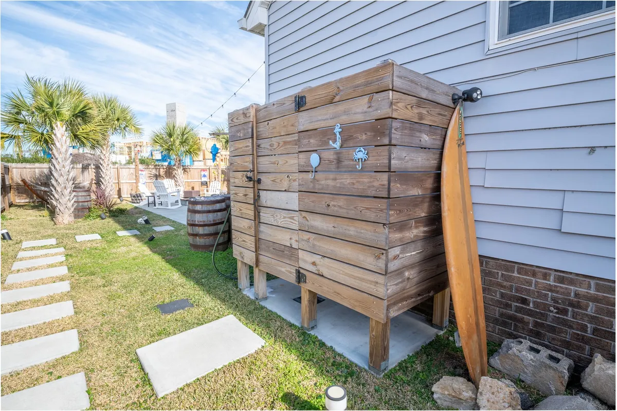 A wooden outdoor shower with a surfboard leaning nearby, set along stepping stones in the backyard. The inviting area is practical for rinsing off after beach days, with lush greenery suggesting a connection to the outdoors and beach lifestyle.