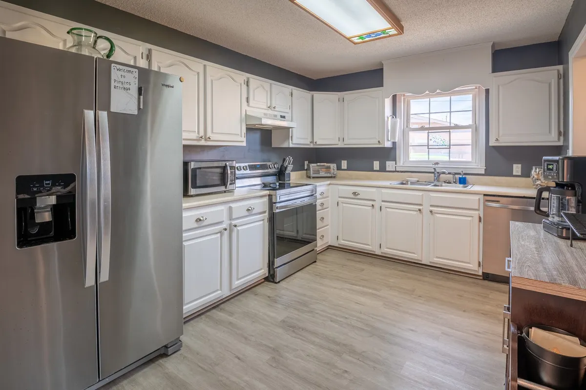 This image showcases a welcoming kitchen space featuring a stainless steel refrigerator, white cabinetry, and kitchen appliances. The room is filled with natural light from a window, emphasizing both functionality and comfort for meal preparation and gatherings.
